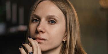 A woman rests her hand near her face while looking toward the camera indoors.