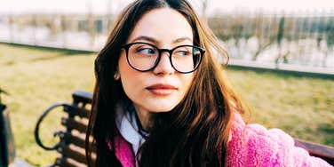 Woman wearing glasses sitting on a park bench and looking off to the side, appearing thoughtful and composed during a quiet moment of reflection.