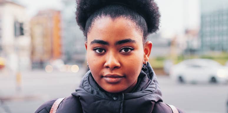 A woman with natural hair looks directly at the camera while standing outdoors in an urban setting.