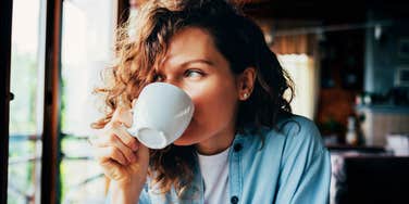 Woman with curly hair drinking coffee by a window in a cozy home setting