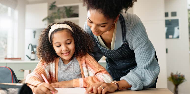 A mother helps her young daughter write in a notebook at a kitchen table.