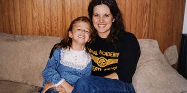 A woman sits on a couch smiling while holding a young child beside her.
