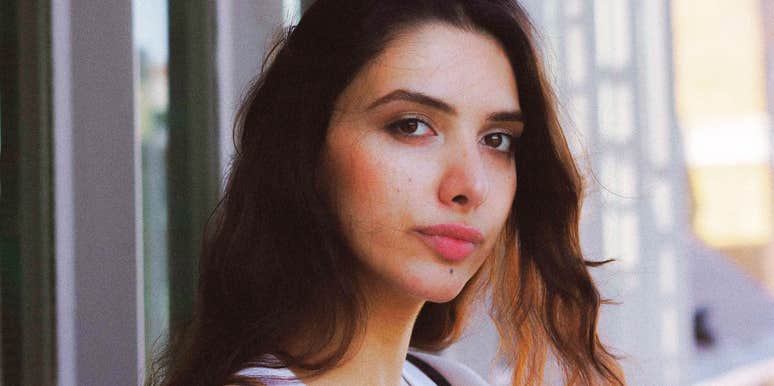 Portrait of a young woman with long dark hair standing near a modern building, looking calmly toward the camera.