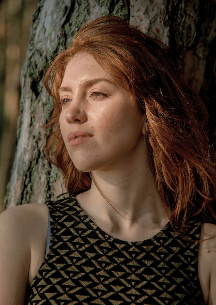 pensive red haired woman leaning against a tree