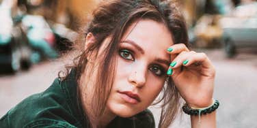A close-up portrait of a young woman with tousled hair resting her hand on her forehead outdoors.
