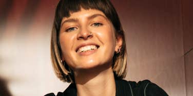 A woman with short hair smiles at the camera indoors under warm lighting.