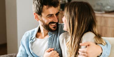 Man smiling warmly at a woman while sitting close together on a couch, sharing a relaxed, affectionate moment