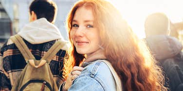 A teenage girl with red hair looking back at the camera while walking outdoors with other people.