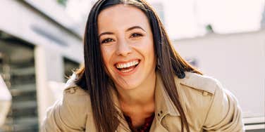 A woman smiles broadly while leaning forward outdoors in daylight.