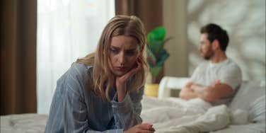 upset woman sitting on edge of bed away from husband